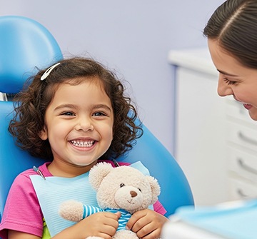Young girl holding a teddy bear and smiling in the dentist’s chair